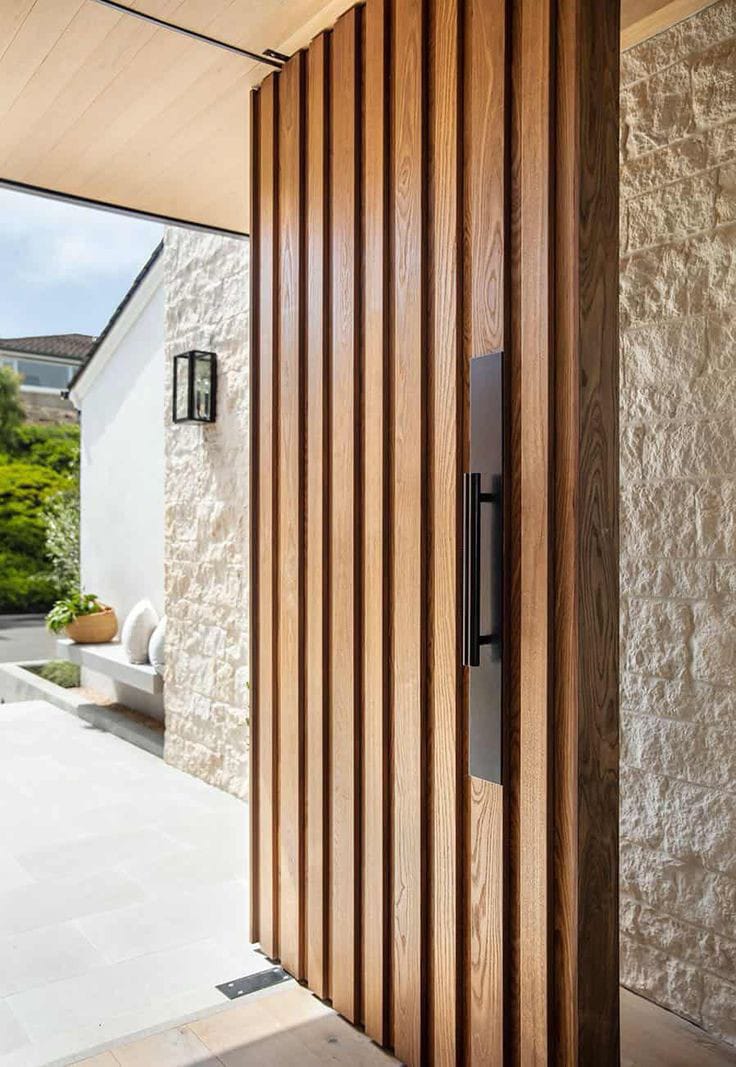 Custom vertical timber-slat pivot entrance door set against light stone cladding